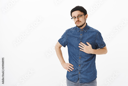 Portrait of bossy confident and handsome skillful male musician in black trendy beanie and shirt pointing at himself with palm on breast squinting and gazing intrigued and doubtful at camera © Cookie Studio