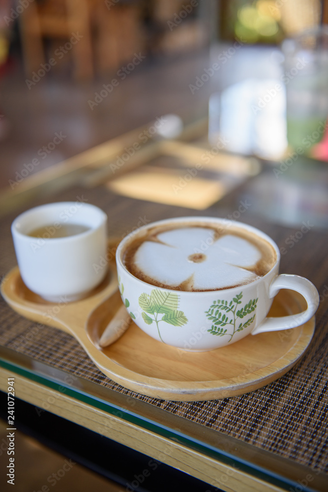 Latte coffee in a white cup, leaf pattern, paired with green tea in a small white cup placed on a wooden plate in a coffee shop. Latte coffee in a white cup, leaf pattern, paired with green tea in a small white cup placed on a wooden plate in a coffee shop.