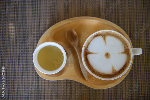 Latte coffee in a white cup, leaf pattern, paired with green tea in a small white cup placed on a wooden plate in a coffee shop.
