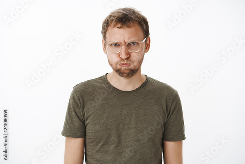 Man feeling mad of friends holding breath and pouting from offence and anger frowning swearing not say any word unless they sorry standing childish and displeased over grey wall in dark-green t-shirt © Cookie Studio