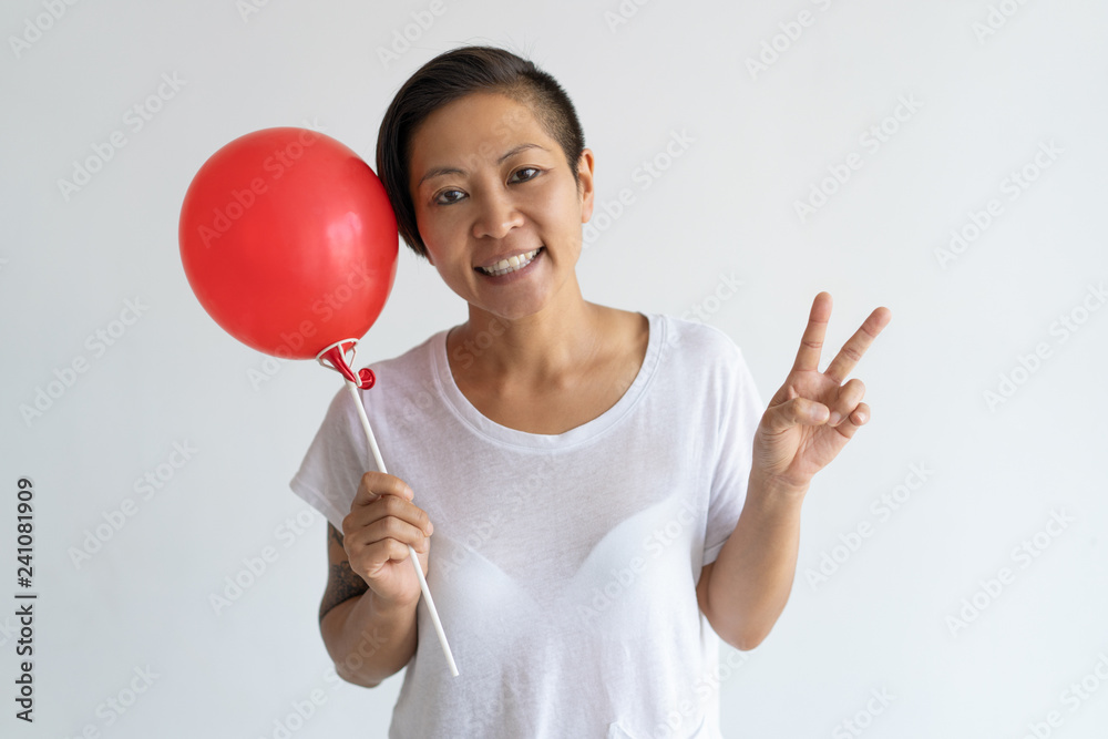Playful Asian woman holding red balloon and showing victory sign. Smiling lady looking at camera. Party concept. Isolated front view on white background.