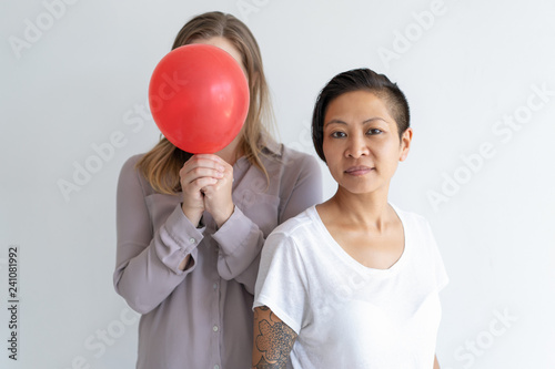 Two women posing at camera and holding red balloon. Multiethnic women standing together. Lesbian couple or women friendship concept. Isolated front view on white background. © Mangostar