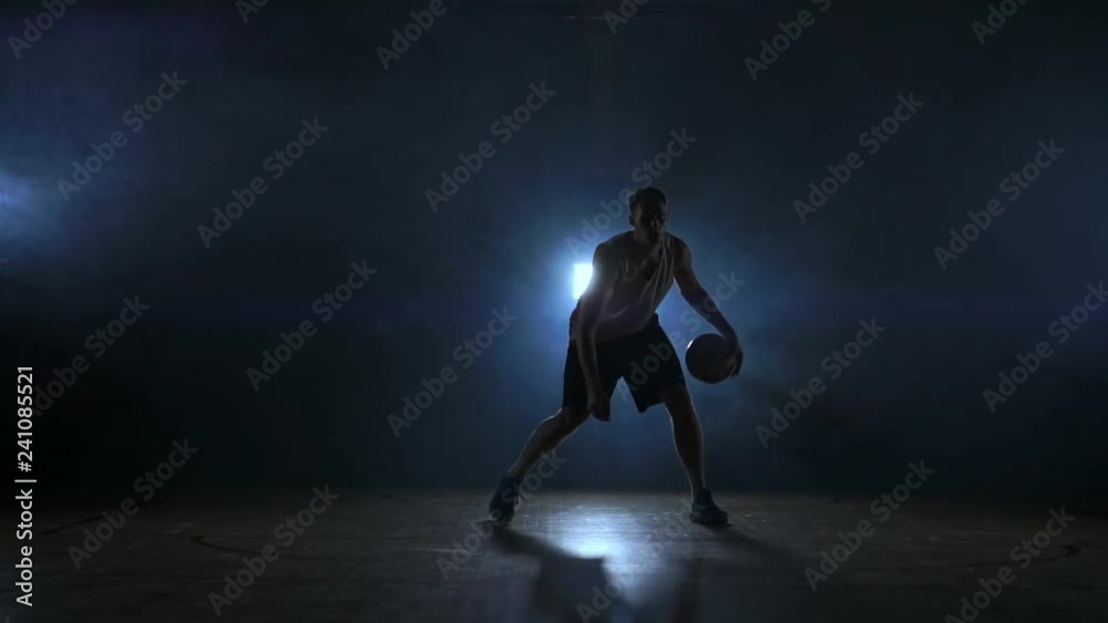 Dribbling basketball player on the court with the ball in a dark room with a backlight in slow motion in the smoke Dribbling basketball player on the court with the ball in a dark room with a backlight in slow motion in the smoke