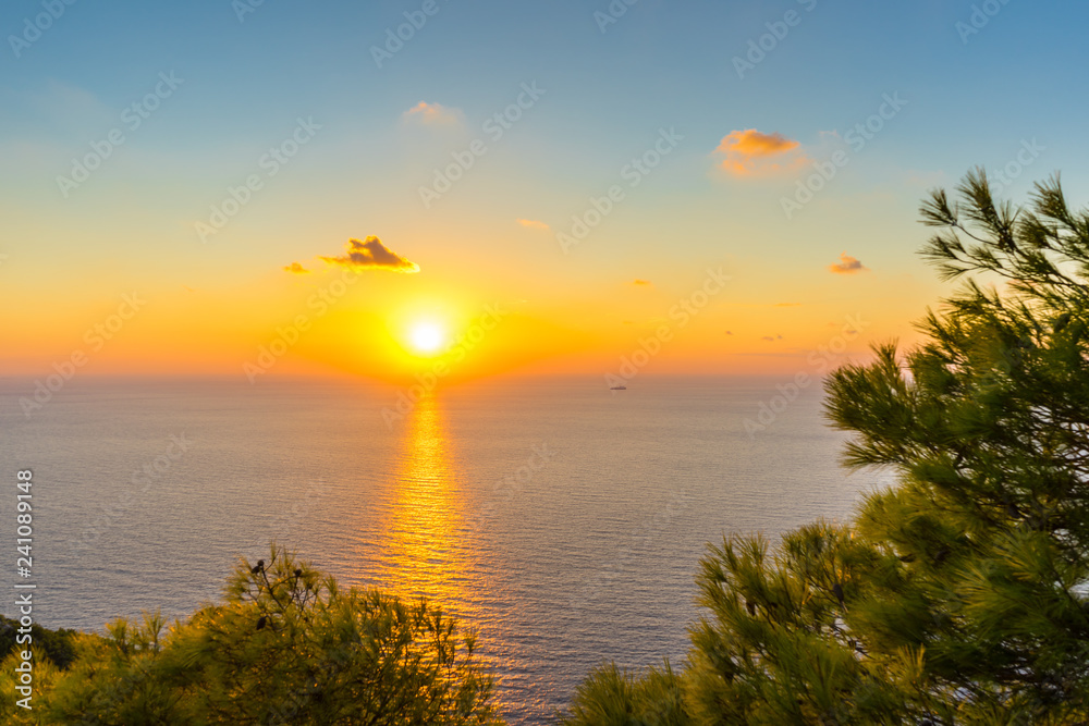 Greece, Zakynthos, Beautiful romantic orange sunset sky reflecting in silent ocean water behind green branches