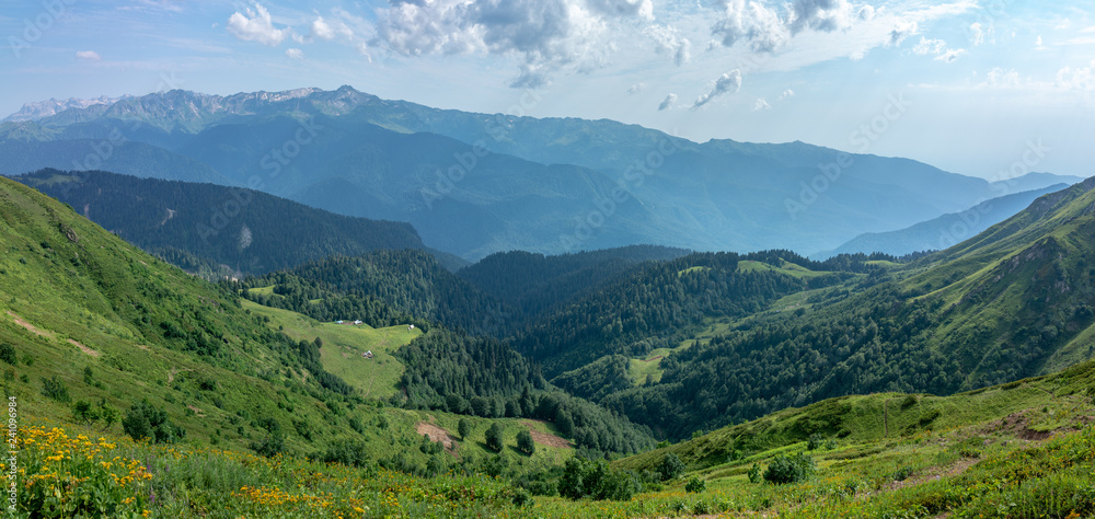 Obraz premium The view from the height of a green mountain valley with residential buildings surrounded by high mountains. Snow-capped mountain peaks on the horizon. Krasnaya Polyana, Sochi, Caucasus, Russia.