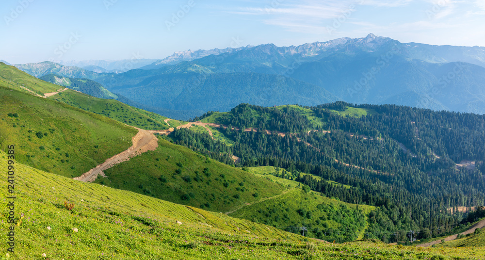 The view from the height of a green mountain valley with residential buildings surrounded by high mountains. Snow-capped mountain peaks on the horizon. Krasnaya Polyana, Sochi, Caucasus, Russia.