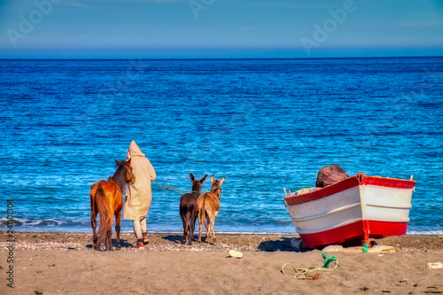 Oued Laou, Chefchaouen, Morocco - November 3, 2018: A young man in traditional Moroccan attire walks his donkeys on the beach of Oued Laou, a small town on the coast of the Mediterranean Sea. Morocco