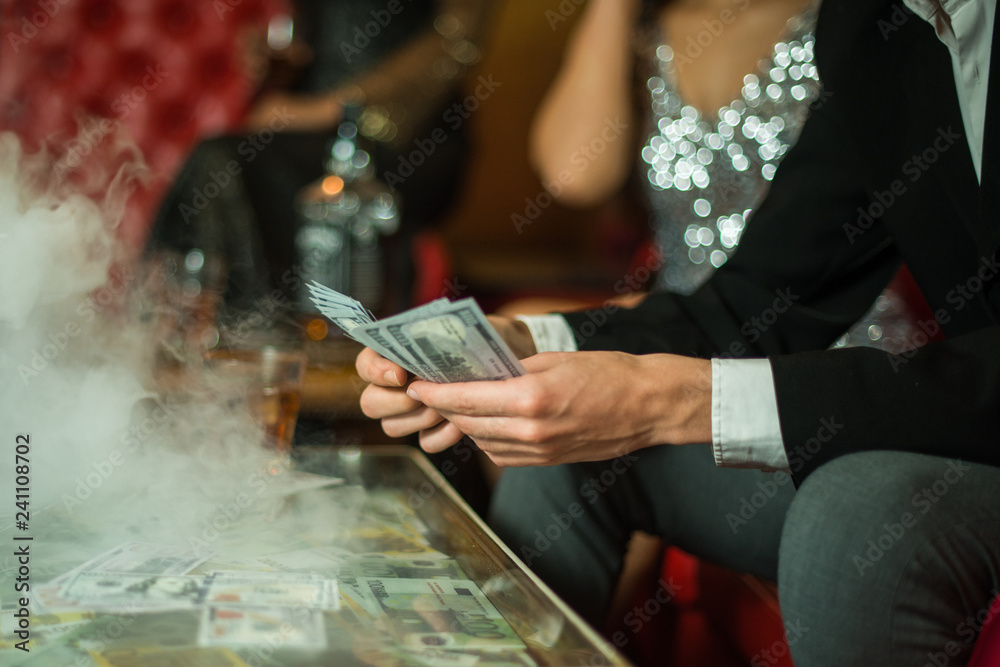 Man with cigar counting money in the club. Group of young multi-ethnic friends relaxing in shisha club-bar in rich interior