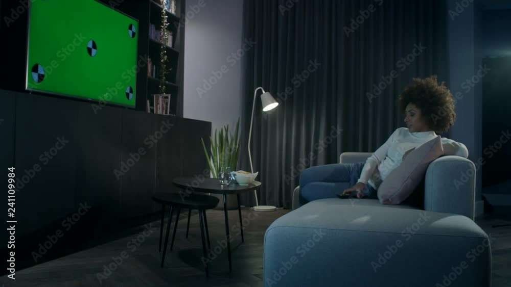 Young woman relaxing on the sofa at night watching a movie on tv. Camera track right to left.