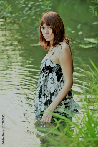 Fototapeta Naklejka Na Ścianę i Meble -  Charming girl bathes in a pond. Beautiful girl in the river near the forest. Young beauty in the water in the hot summer. Sexy brown hair relaxes near the river. Mysterious Amazon.