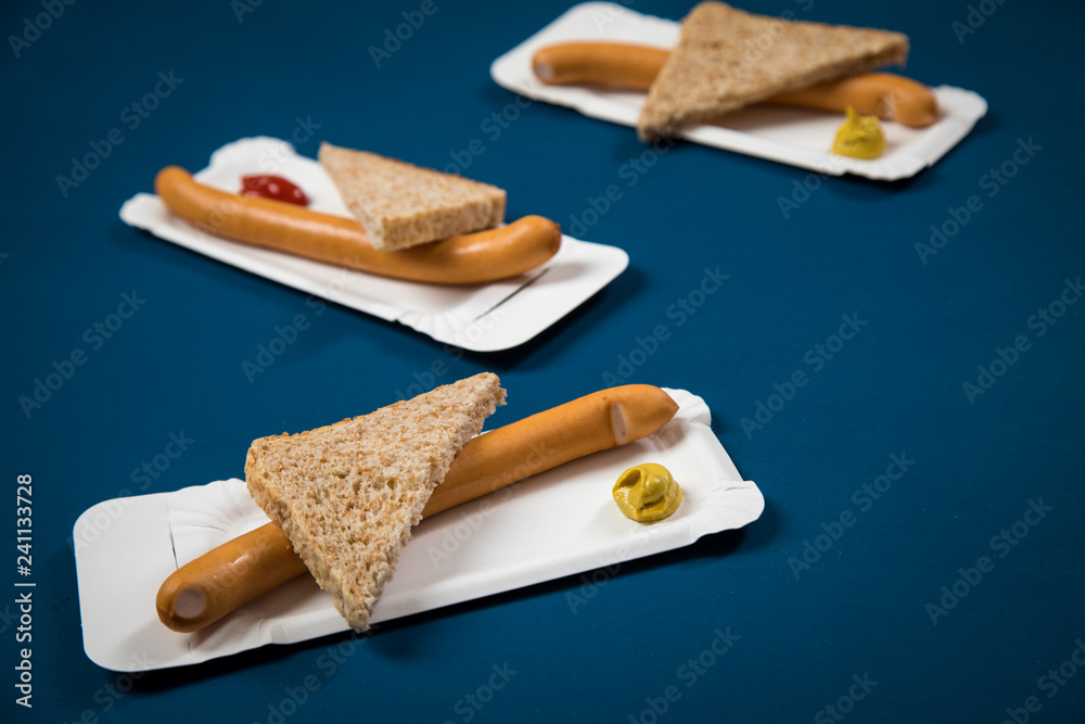 Fototapeta premium Wiener Würstchen mit Vollkorn Brot Scheibe auf Einweg Pappteller mit Senf und Ketchup auf Hintergrund Blau