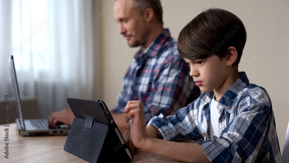 Male kid playing game on tablet while father working on laptop at home, gadget Male kid playing game on tablet while father working on laptop at home, gadget