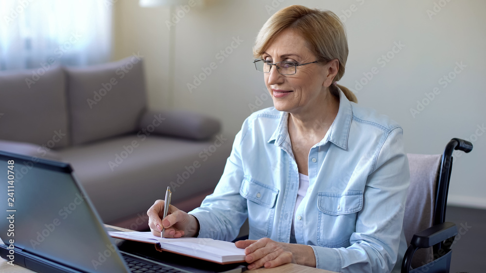 Senior woman in wheelchair studying online and making notes, video lessons Senior woman in wheelchair studying online and making notes, video lessons