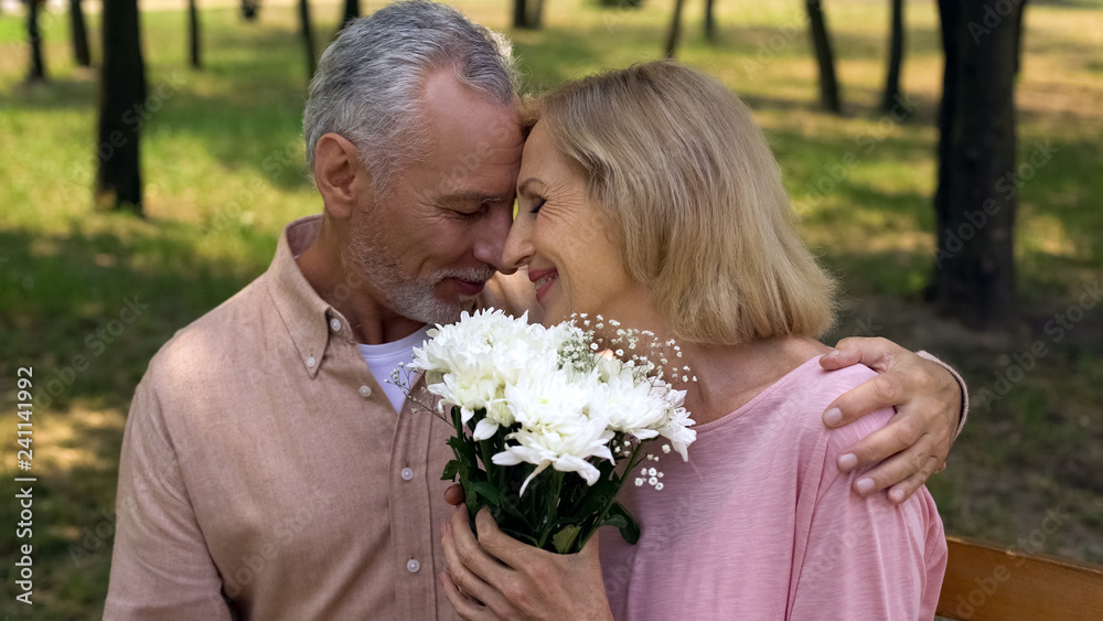 Loving aged couple nuzzling during romantic date in park, flowers bunch present Loving aged couple nuzzling during romantic date in park, flowers bunch present