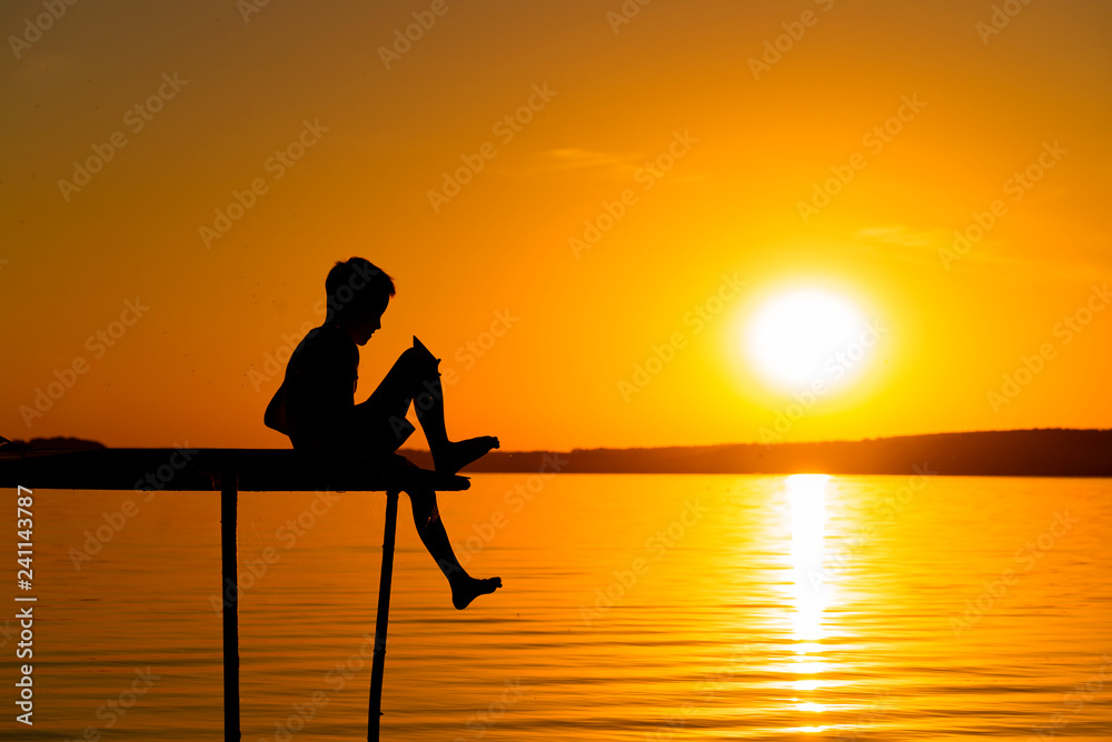 Silhouette of a happy boy sitting on the end of a bridge over the river at sunset. A small boy sits alone on a wooden bridge on the background of a river in the evening.