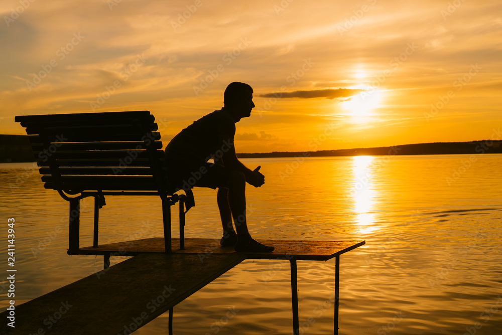 Obraz premium Single man sitting on the bench on a small bridge in the evening and looking at sunset. View of a man sitting and resting alone on wooden bench at river, lake or sea.