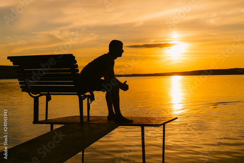 Single man sitting on the bench on a small bridge in the evening and looking at sunset. View of a man sitting and resting alone on wooden bench at river, lake or sea.