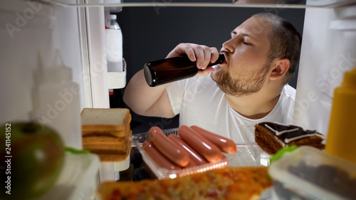 Fat man drinking beer with pleasure from fridge at night, unhealthy lifestyle