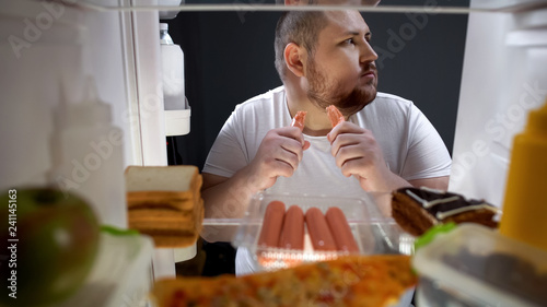 Addicted man secretly eating sausages at night near fridge, unhealthy nutrition