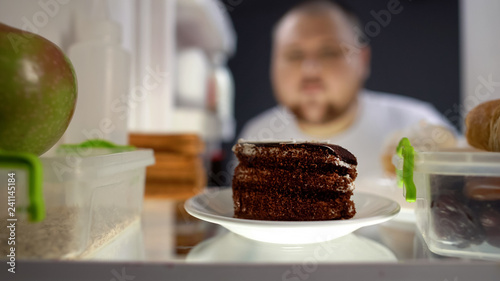 Obraz na plátne Oversize man taking piece of cake from fridge at night, diabetes risk, calories
