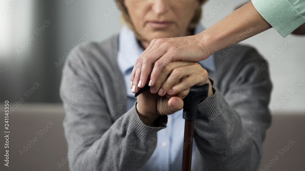 Elderly retired lady sitting on sofa, young woman touching her hands carefully