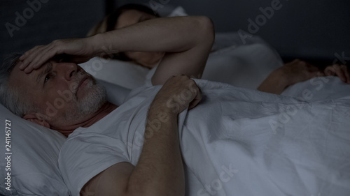 Elderly male lying sleepless in bed suffering headache, rubbing head, troubles