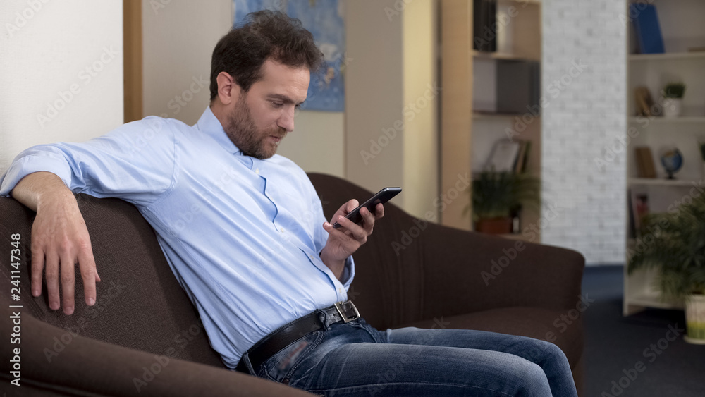 Man on couch reading news on smartphone, using mobile banking application