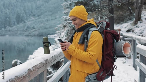 Young traveler man using smartphone in hiking winter tour. Snowy blurred mountains cape and lake. Traveling and communication concept: hiker scrolling and tapping on line by cell phone photo