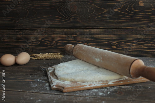 Тесто на деревянной доске посыпанное мукой , яйца и колоски пшеницы на столе.
