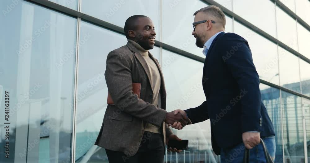 African American young man in casual style standing at the modern glass building and chatting on the phone, then his colleague businessman coming, shaking hands and talking. Outside. African American young man in casual style standing at the modern glass building and chatting on the phone, then his colleague businessman coming, shaking hands and talking. Outside.