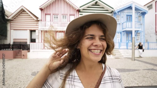 Cute young female smiling and running on camera near colorful houses. POV view portrait happy girl in hat. Concept tourist have fun. Costa Nova, Portugal