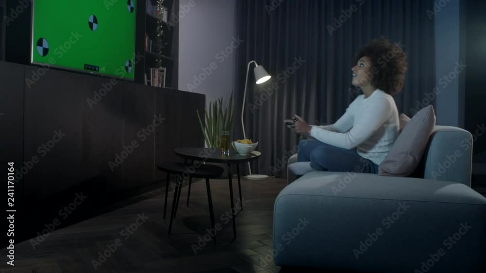 A competitive woman sitting cross legged on a sofa at home at night playing a computer game on a large flat screen tv.