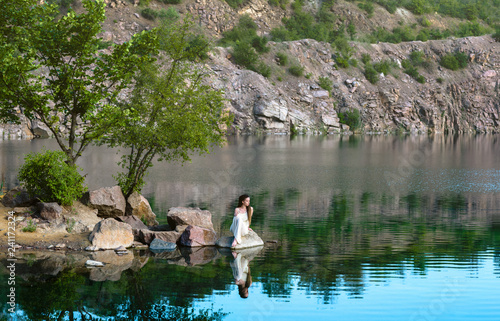 Beautiful girl sitting on a rock in a light dress. On the shore of the lake.