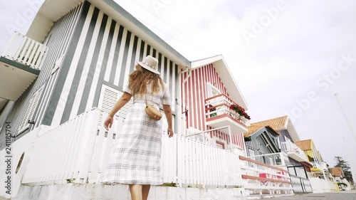 Playful woman whirling and dancing on street of fisherman village Costa Nova. Bottom wide angle shot flapping dress and raised hands. Girl at background colorful houses and bright sky. Portugal