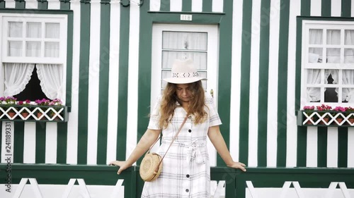 Cozy house on street Costa Nova village. Woman in hat standing near fence looking at camera and smiling on background facade with door and cute windows. Costa Nova, Portugal