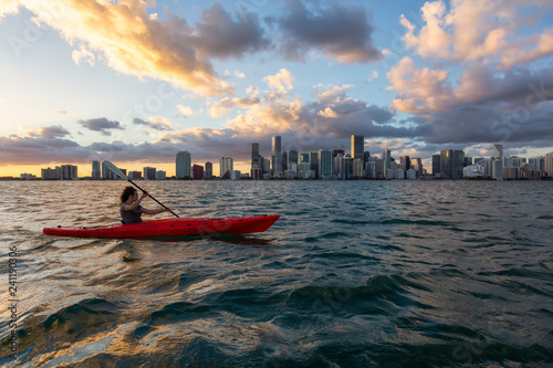 Adventurous girl kayaking in front of a modern Downtown Cityscape during a dramatic sunset. Taken in Miami, Florida, United States of America.