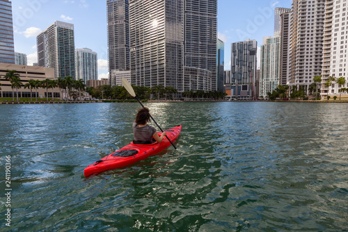 Adventurous girl kayaking in front of a modern Downtown Cityscape during a sunny evening. Taken in Miami, Florida, United States of America.