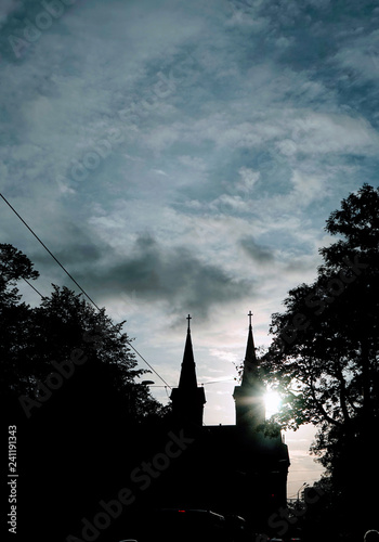 In Tallinn Estonia trees frame silhouetted steeples against a stormy sky with the sun glistening behind a steeple..