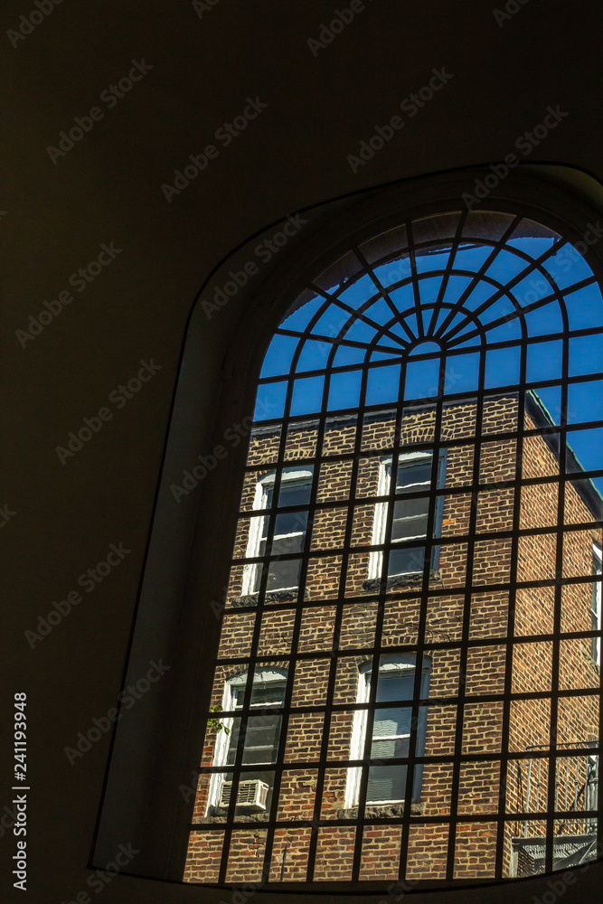View a brownstone building through a large vertical multi-paned window with a curved top. The brownstone has four white framed windows and fire escape. Blue sky is above the building.