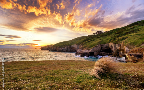 An epic sunset with dramatic burning clouds in Tunnel Beach of Dunedin, New Zealand. One can enjoy ocean view, cliff, rolling hills, sunset, coves, rock formation in this famous tourist attraction.