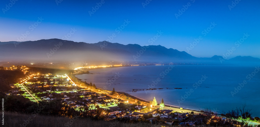 The town of Kaikoura, New Zealand. This was shot from a hill. It was dusk and the sky turns deep blue. The city lights are bright. This picturesque coastal town is the perfect place for travelers.