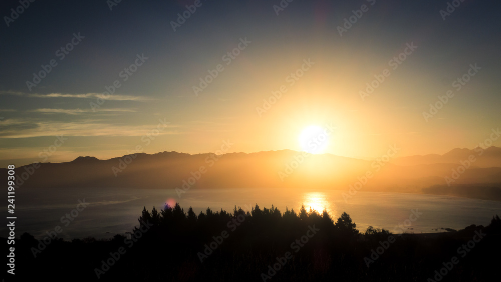 A brand new day as the sun rises above the mountain. Below are ocean and tree line. This image is so beautiful. It has a peaceful, serene and calming feel to it. It is suitable for background use.