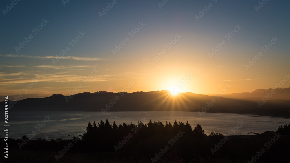 A brand new day as the sun rises above the mountain. Below are ocean and tree line. This image is so beautiful. It has a peaceful, serene and calming feel to it. It is suitable for background use.