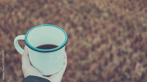 hand holding hot black coffee in white melamine cup and blurred brown field as background