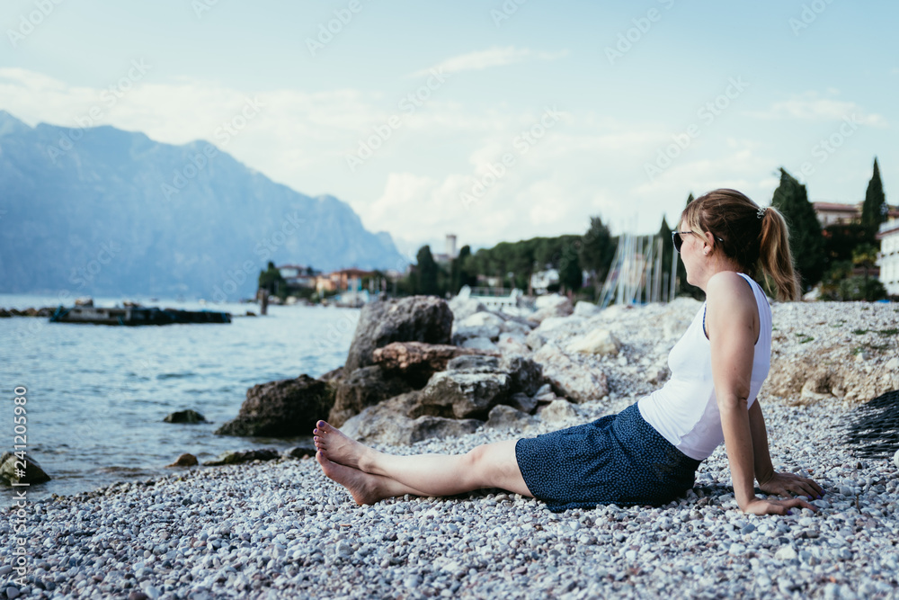 Beautiful young woman is sitting on the pebble beach and enjoying the view, vacation
