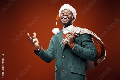 Modern Santa Claus. Smiling emotional man posing in green coat and red sweater, with santa hat and bag. Studio shot, brown background