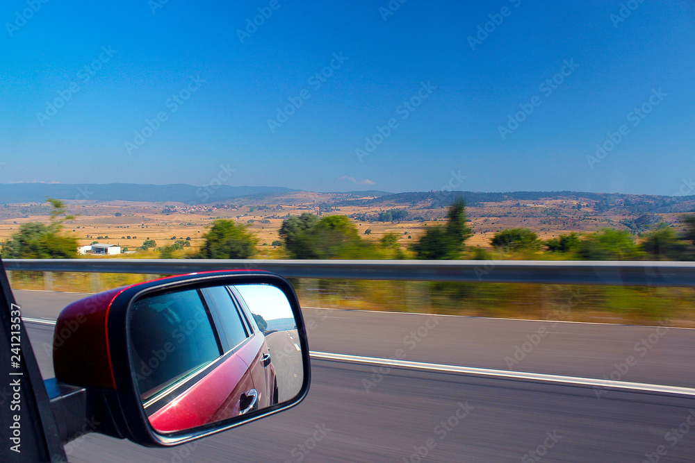Red SUV driving at speed on the road. In the rear view mirror displays the roadbed with the markup and the car itself. View of the mountain landscape from the car window.