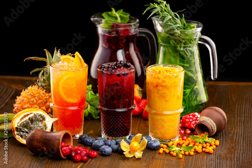 Three glasses of homemade healthy lemonades with tarragon, mint, citrus mix, black currant and sea-buckthorn at wooden table decorated with fruits background.