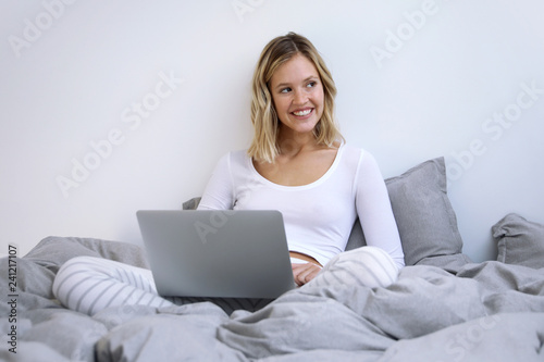 Frau mit Laptop sitzt lachend auf einem Bett