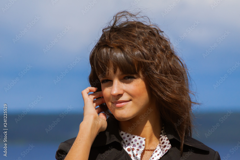 closeup portrait of beautiful smiling woman in black jacket, with long dark brown curly hair outdoor at sunny windy day, on blue sky background looking into the camera